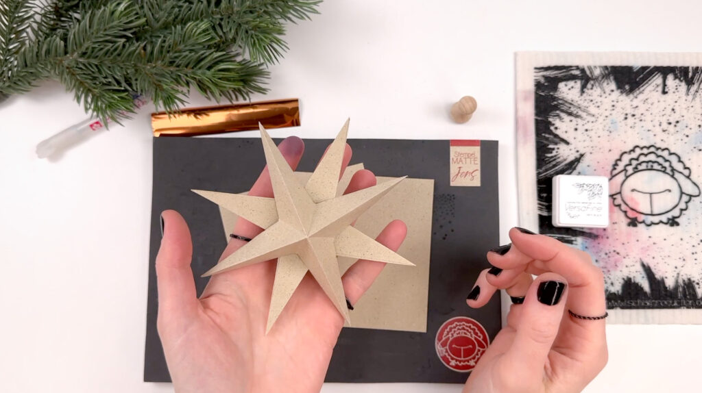 Eine Hand präsentiert einen selbstgebastelten Adventsstern aus Graspapier, im Hintergrund Tannenzweig, schwarze Stempelmatte, Stempel, Stempelkissen und goldene Transferfolie.