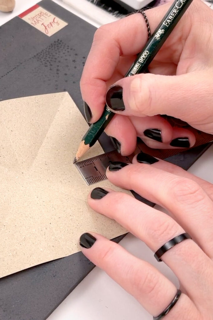 Graspapier mit Knicke auf einer schwarzen Stempelmatte, eine Hand legt ein Lineal an, während die andere einen Bleistift an die Linealkante hält.