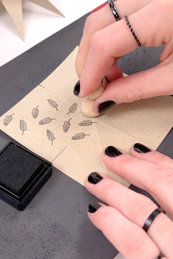 Graspapier mit Schnittkanten auf einer schwarzen Stempelmatte, eine Hand hält das Papier, während die andere mit einem Holzstempel Tannenzweig-Motive stempelt, nebenbei liegt ein schwarzes Stempelkissen.