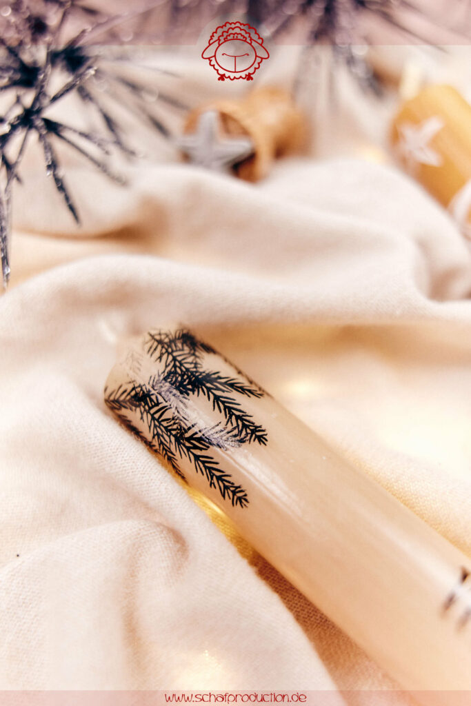 Zwei Stabkerzen auf cremefarbener Decke, eine cremefarben mit schwarzen Tannenzweigstempeln und 'Weihnachtszauber'-Schriftzug, die andere golden mit weißen Tannenzweigstempeln und 'Weihnachtszauber'-Schriftzug, umgeben von Stempeln und silbernen Tannenzweigen im Hintergrund.