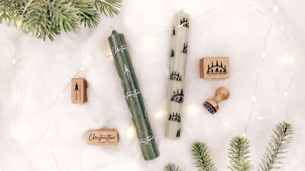 Zwei dekorativ bestempelte Stabkerzen auf weißem Untergrund. Eine grüne Kerze mit weißen Tannenbaumstempeln und dem Schriftzug ‚Christmastime', die andere hell mit schwarzen Schwibbogenstempeln. Im Hintergrund sind Stempel, grüne Tannenzweige und eine feine Lichterkette zu sehen.