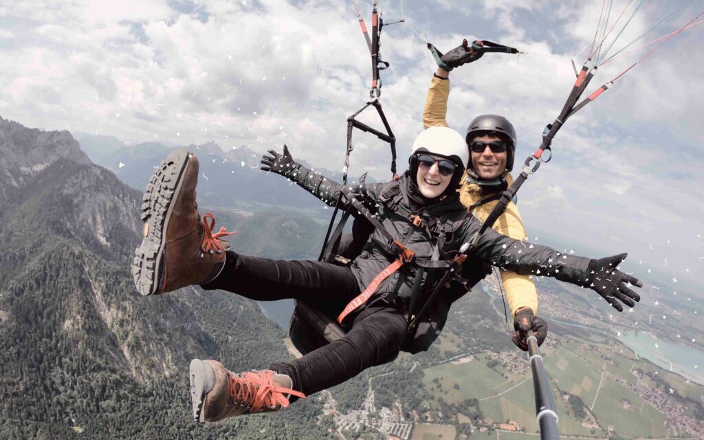 Auf dem Bild sieht man zwei Personen beim Paragliding. Im Hintergrund sind Berge