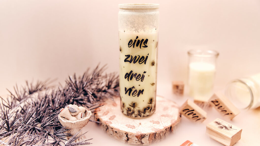 Adventskerze in einem Glas auf einem Holzbrett, beschriftet mit 'Eins, Zwei, Drei, Vier', umgeben von Stempeln, Bastelmaterialien und silbernen Tannenzweigen im Hintergrund.