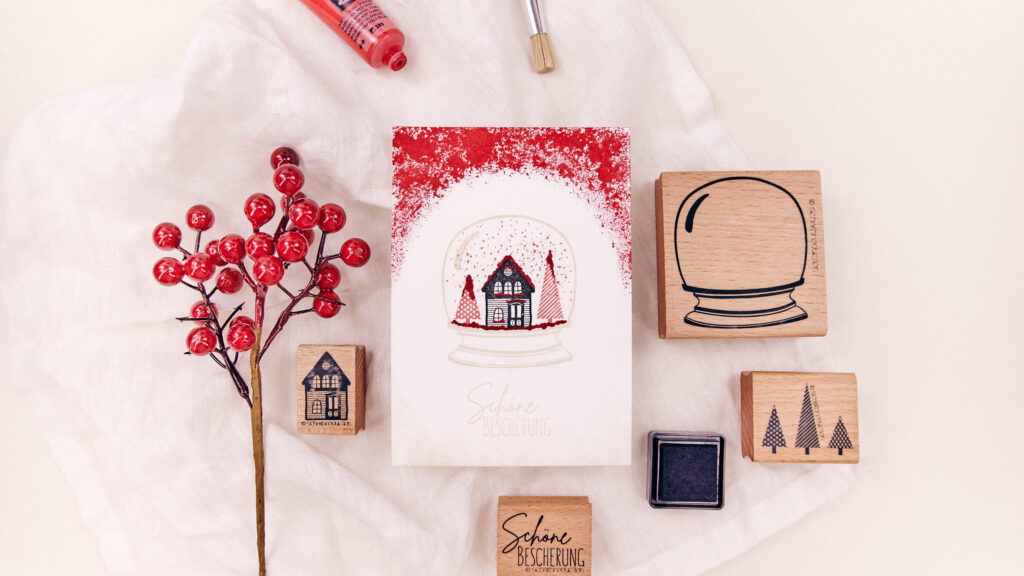 Handgefertigte Postkarte mit Schneekugel-Motiv, in der ein Haus unter rotem Schnee und Tannenbäume dargestellt sind, umgeben von Bastelmaterialien und Stempeln.