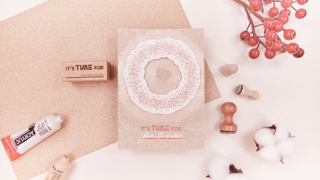 Handgemachte Weihnachtskarte aus Graspapier mit einem Kranz aus Mistelzweigen und Sternmotiven in Rot, Weiß und Gold, darunter der Schriftzug 'its time for love and kisses'.